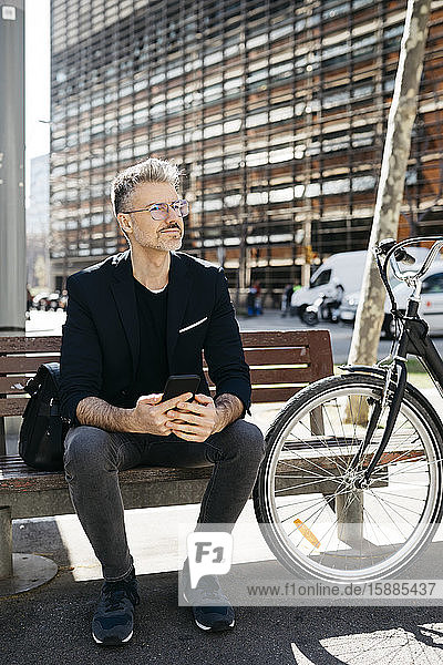 Grauhaariger Geschäftsmann  der in der Stadt auf einer Bank neben dem Fahrrad sitzt