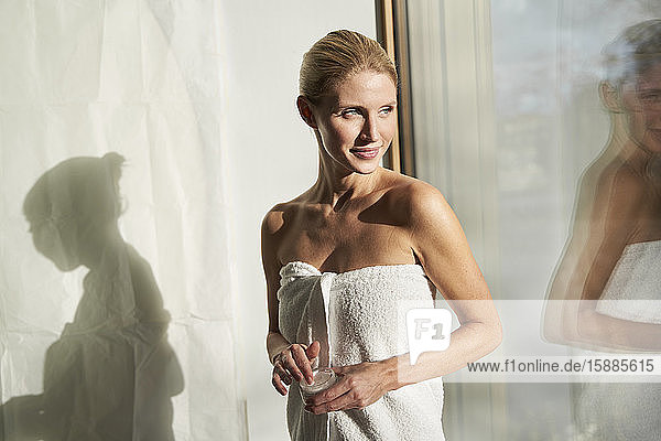 In ein Handtuch gewickelte Frau steht am Fenster und hält einen Sahnebehälter