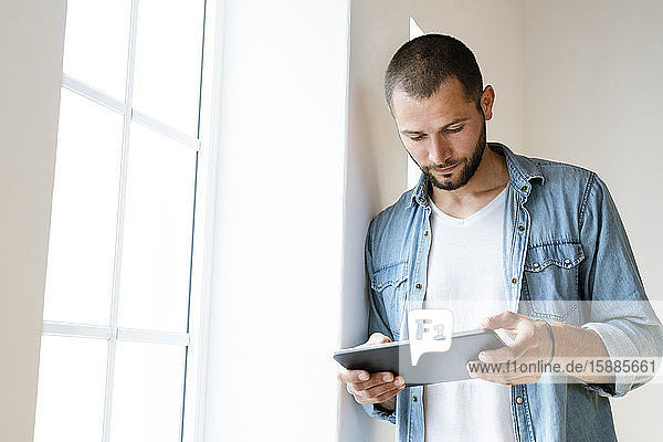 Young serious man at home looking at his tablet standing at window