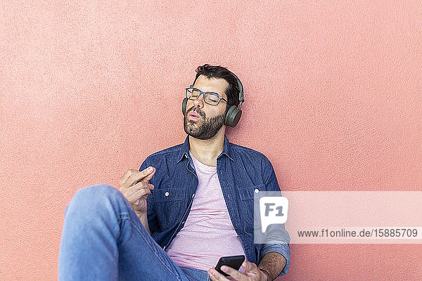 Porträt eines pfeifenden Mannes mit geschlossenen Augen  der sich an eine rosa Wand lehnt und mit Kopfhörern Musik hört