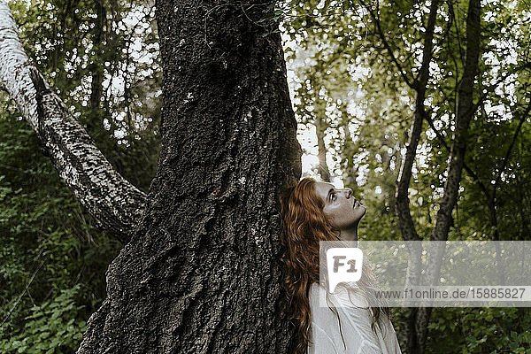 Junge rothaarige Frau umarmt Baumstamm im Wald