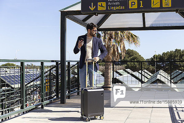 Businessman with smartphone and trolley bag standing on roof terrace of an airport
