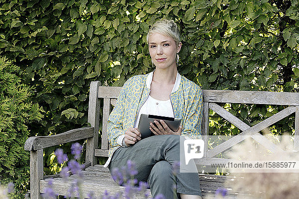 Porträt einer Frau mit digitalem Tablett  die sich auf einer Holzbank in einem Park entspannt