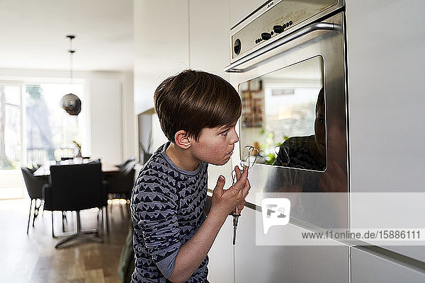 Junge schmeckt Teig,  während er sein Spiegelbild auf der Oberfläche des Ofens betrachtet
