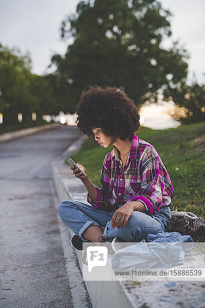 Junge Frau mit Afro-Frisur sitzt mit Smartphone auf dem Bordstein