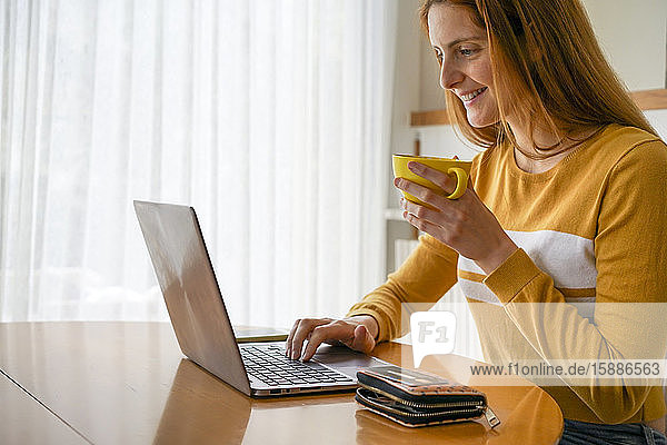 Smiling young woman with cup of coffee using laptop at home