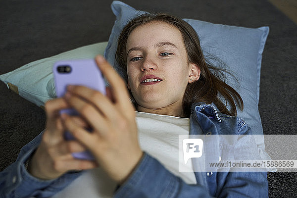 Porträt eines Mädchens  das zu Hause auf dem Boden liegt und auf ein Smartphone schaut