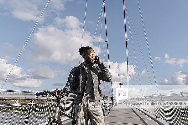 Stylischer Mann mit Fahrrad beim Telefonieren auf einer Brücke