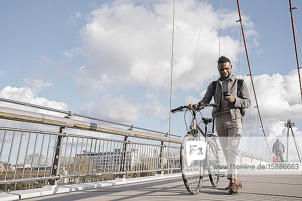 Stylischer Mann mit Fahrrad und Smartphone beim Gehen auf einer Brücke