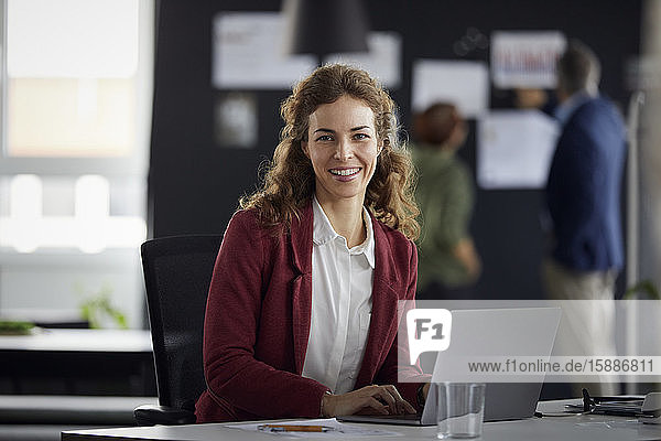 Portrait einer lächelnden Geschäftsfrau mit Laptop am Schreibtisch im Büro mit Kollegen im Hintergrund