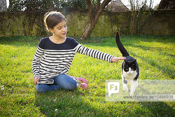 Porträt eines kleinen Mädchens auf einer Wiese sitzend mit Mini-Tablette streichelnder Katze