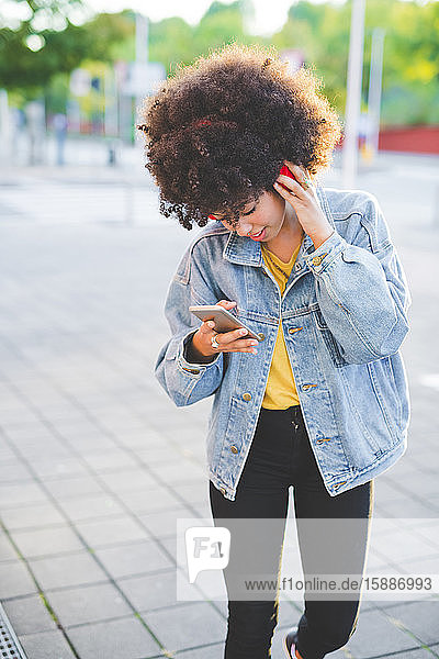 Junge Frau mit Afrofrisur benutzt Smartphone und Kopfhörer in der Stadt