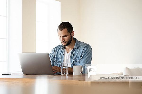 Fokussierter junger Mann arbeitet zu Hause am Laptop im Home-Office im modernen Wohnzimmer