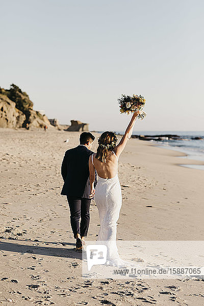 Rückansicht eines glücklichen Brautpaares beim Spaziergang am Strand