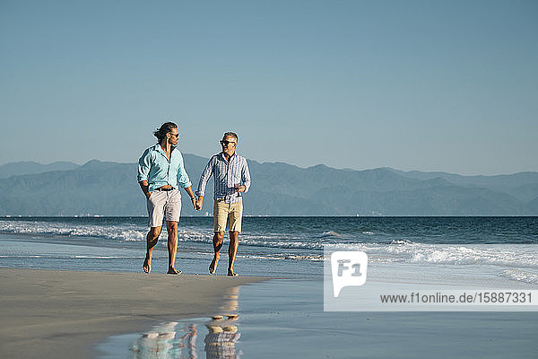 Reife schwule Männer in voller Länge halten sich an den Händen  während sie am Strand vor klarem blauen Himmel an der Küste spazieren gehen  Riviera Nayarit  Mexiko