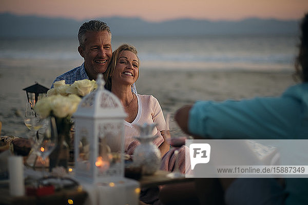Lächelndes reifes Paar  das einen männlichen Freund ansieht  während es am Strand der Riviera Nayarit  Mexiko  ein Abendessen bei Sonnenuntergang genießt.