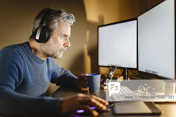 Älterer Mann mit Kopfhörern sitzt zu Hause am Schreibtisch und arbeitet am Computer