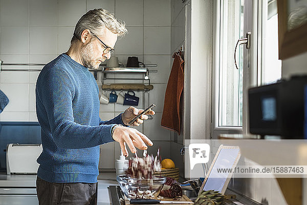 Reifer Mann steht in seiner Küche und schaut auf sein Smartphone  während er Salat zubereitet