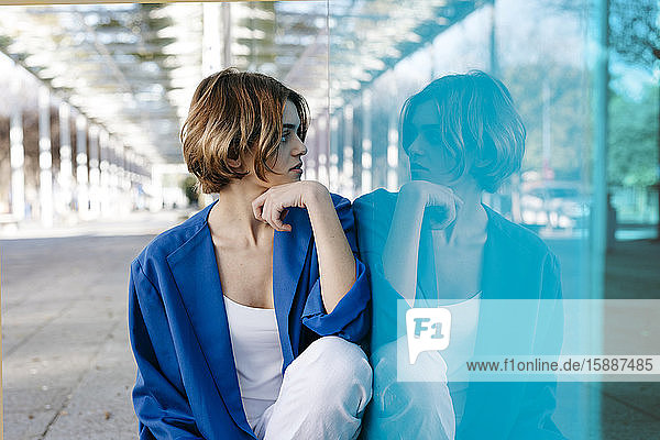 Junge Frau mit urbanem Aussehen sitzt auf dem Boden und lehnt mit ihrem Spiegelbild an einer bunten Glaswand