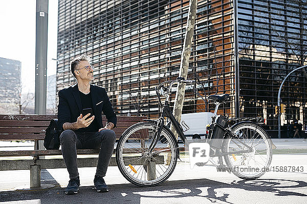 Gray-haired businessman sitting on a bench next to electric bicycle in the city