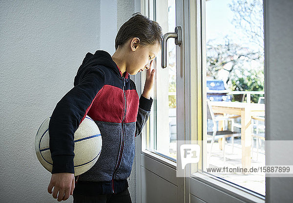 Trauriger Junge mit an das Fenster gelehntem Basketball