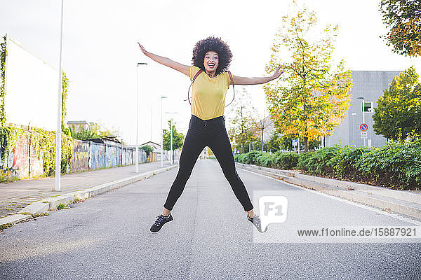 Glückliche junge Frau mit Afrofrisur springt auf einer Straße in der Stadt