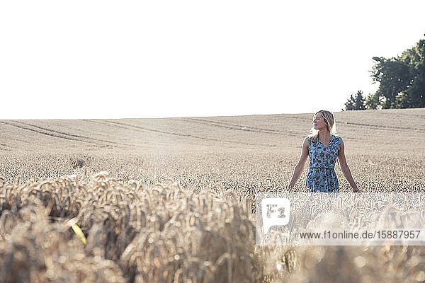 Blonde Frau steht bei Sonnenuntergang in einem Weizenfeld und schaut in die Ferne