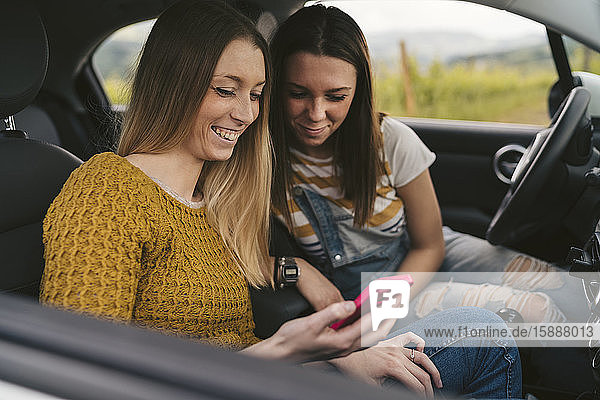 Zwei glückliche junge Frauen auf einer Autoreise  die sich ein Handy teilen