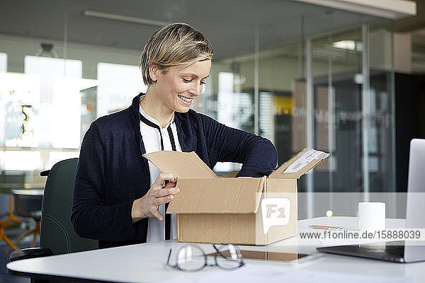 Lächelnde Geschäftsfrau packt Paket auf Schreibtisch im Büro aus