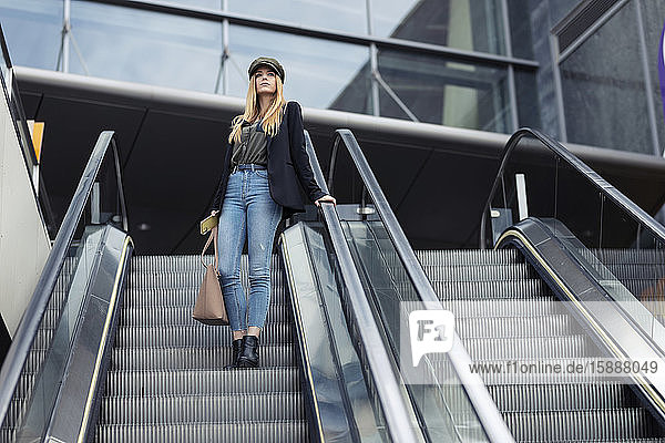 Blonde junge Frau mit Tasche und Smartphone steht auf einer Rolltreppe und schaut in die Ferne