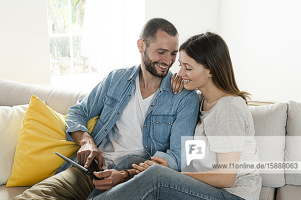 Happy couple at home in modern living room sitting on couch while looking at tablet together