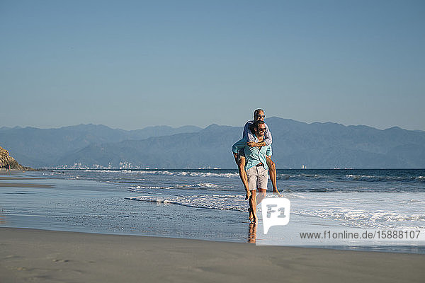 Reifer schwuler Mann nimmt seinen Partner huckepack  während er am Strand bei strahlend blauem Himmel an der Küste spazieren geht  Riviera Nayarit  Mexiko