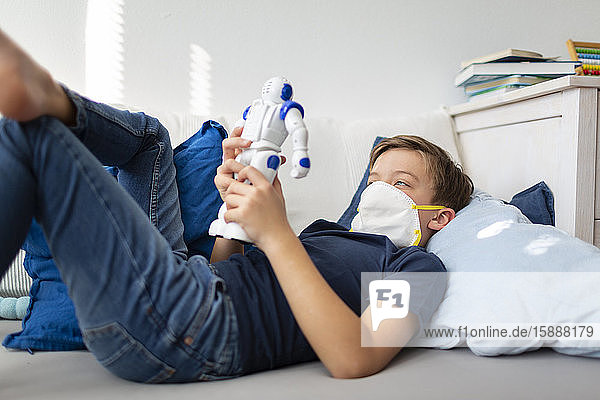 Junge mit Maske spielt während der Koronakrise mit seinem neuen einzigen Freund einem Roboter in seinem Zimmer