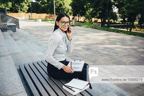 Lächelnde Geschäftsfrau ruft an und sitzt auf einer Bank in einem Park