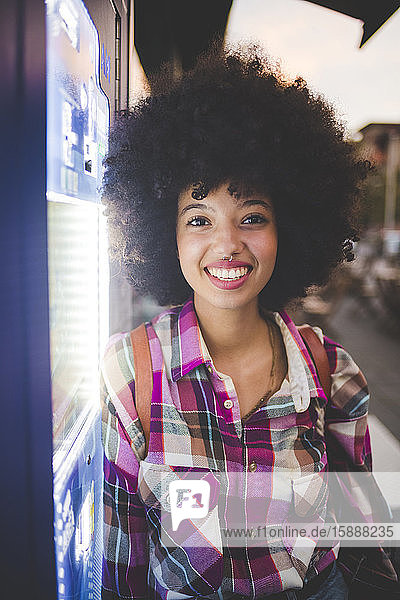Portrait of happy young woman with afro hairdo leaning against vending machine in the city at dusk