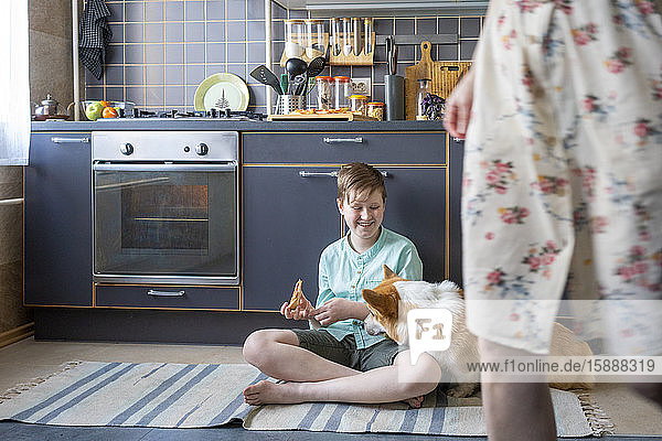 Lächelnder Junge sitzt mit einem Stück Pizza auf dem Küchenboden und schaut den Hund an