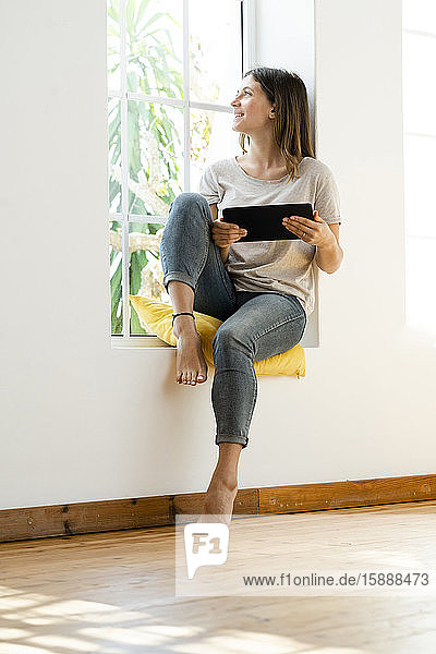 Smiling young brunette woman at home sitting on window bench and holding her tablet looking outside