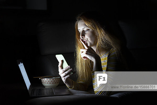 Junge Frau liegt zu Hause auf der Couch und benutzt Laptop und Smartphone