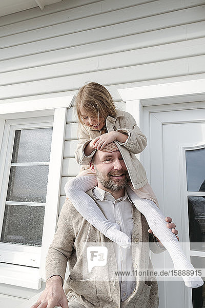 Porträt eines Vaters mit glücklicher Tochter auf den Schultern vor ihrem Haus