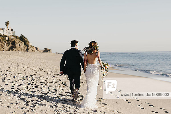 Rückansicht des am Strand laufenden glücklichen Brautpaares