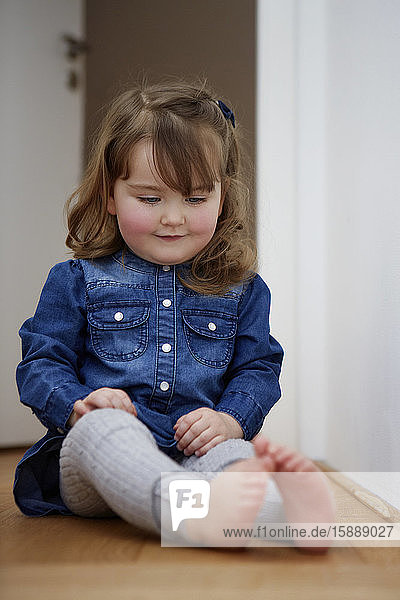Porträt eines lächelnden Kleinkindes  das zu Hause auf dem Boden sitzt und auf ihre nackten Füße schaut