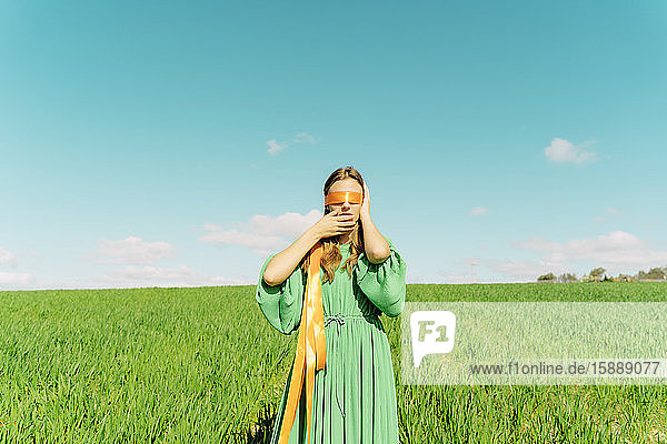 Junge Frau mit verbundenen Augen  die ein grünes Kleid trägt und in einem Feld steht