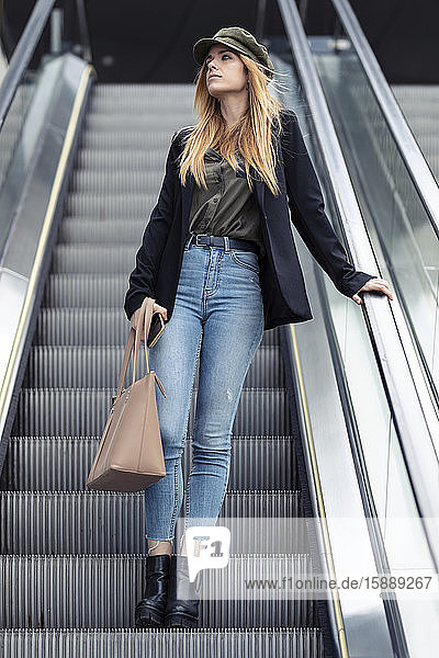Porträt einer blonden jungen Frau mit Tasche und Smartphone auf einer Rolltreppe stehend