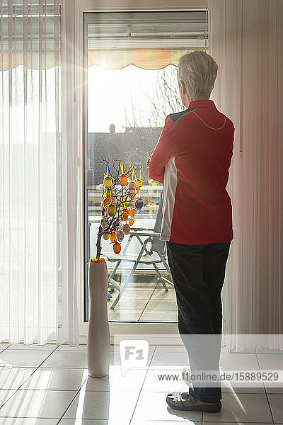 Ältere Frau steht auf geschlossenem Balkon und schaut aus dem Fenster