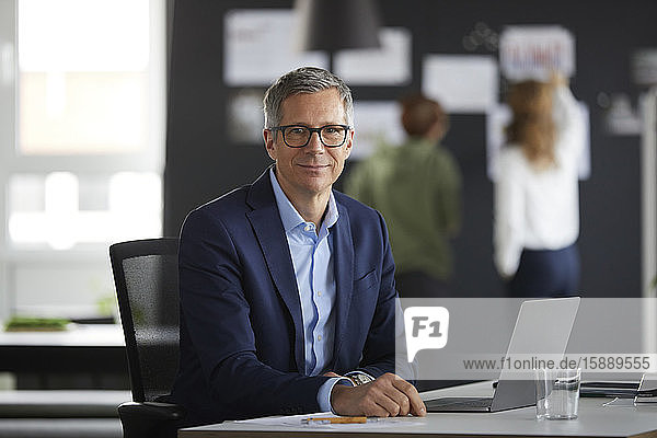 Porträt eines Geschäftsmannes am Schreibtisch im Büro mit Kollegen im Hintergrund