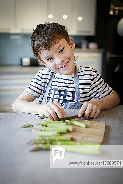 Porträt eines lächelnden kleinen Jungen  der in der Küche grünen Spargel schneidet