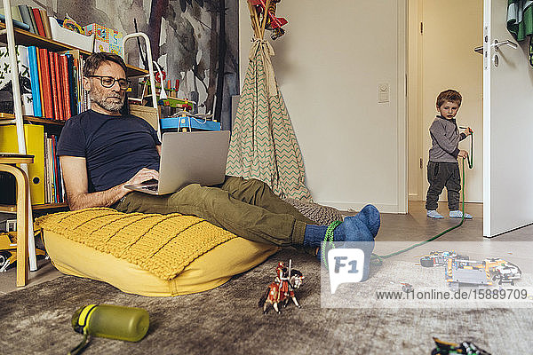 Son tying his father working on laptop in children's room