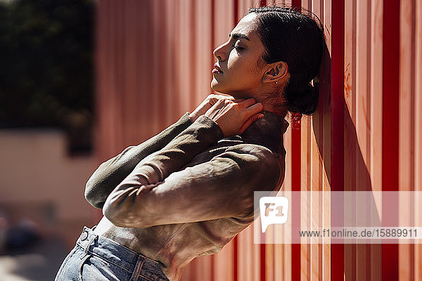 Side view of beautiful young woman leaning on red corrugated wall during sunny day