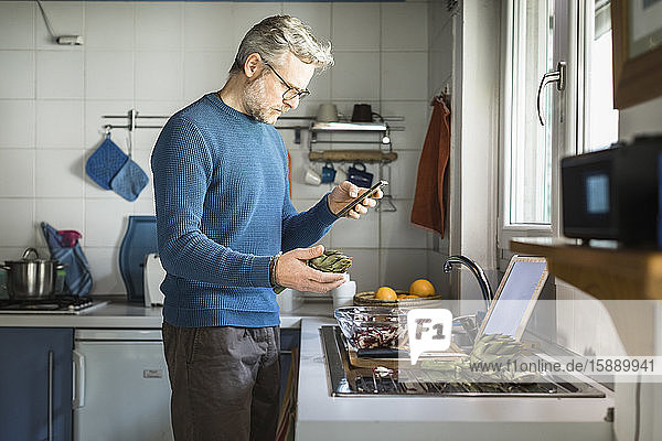 Reifer Mann  der in seiner Küche Artischocken zubereitet und auf sein Smartphone schaut