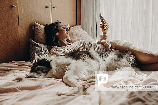 Frau entspannt sich mit ihrem Hund im Bett und schaut auf Smartphone